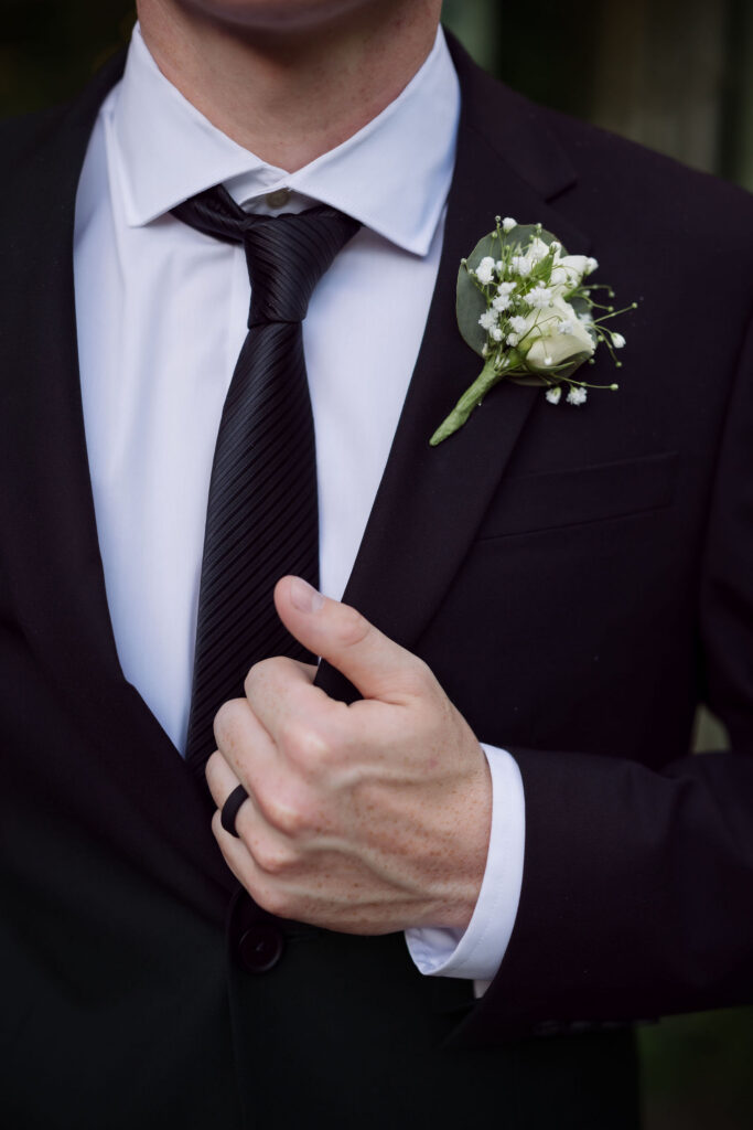 Close-up of the groom’s white rose and baby’s breath boutonniere, black tie, and wedding band at one of the rustic Gatlinburg chapels.