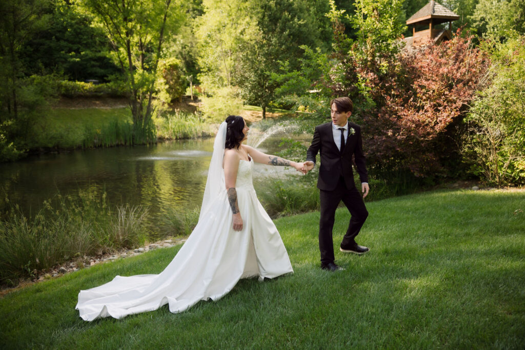 Gatlinbugr elopement at little log chapel in the smokies with bride and groom holding hands as the groom leads the bride through a small lawn in front of the lake
