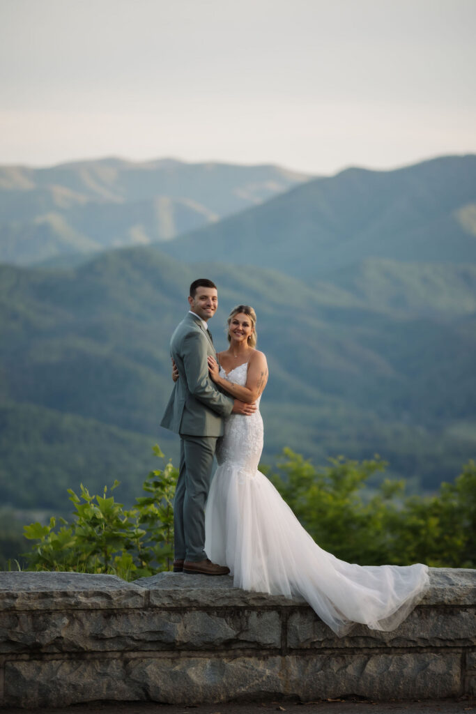 Bride and groom pose together on a stone overlook with layered Smoky Mountain views behind them.