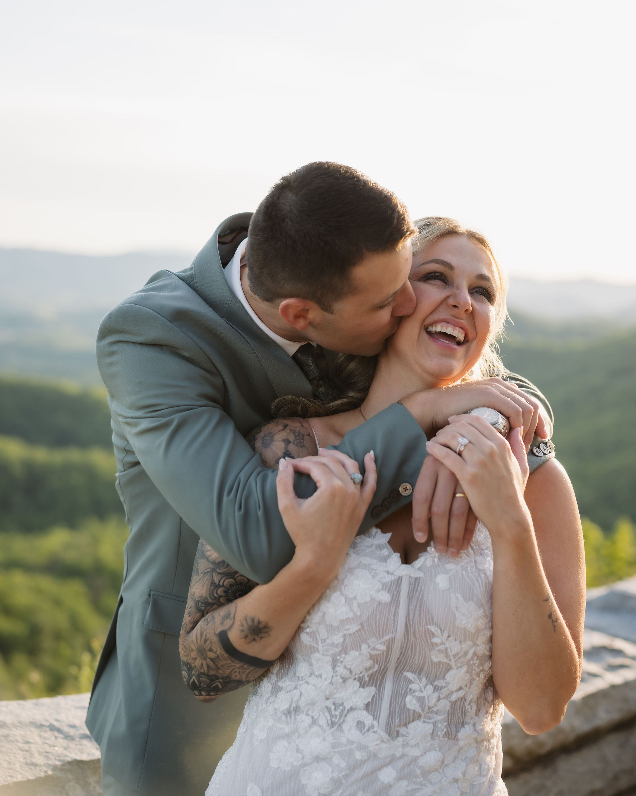 Groom kisses the bride’s cheek as they embrace at Foothills Parkway, with soft mountain views in the background during their wedding portraits.