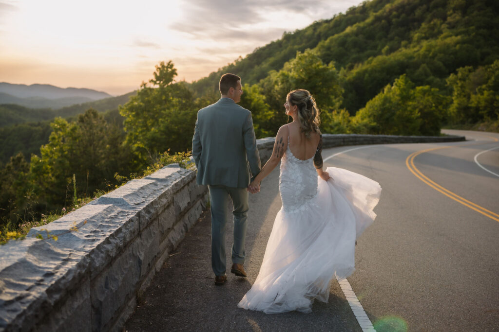 Bride and groom walk hand in hand along a mountain road at sunset during their Smoky Mountain wedding.