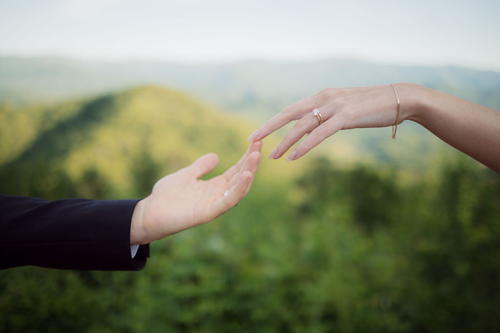 Bride and groom reach toward each other with wedding rings visible against the mountain scenery in creative Foothills Parkway photos.