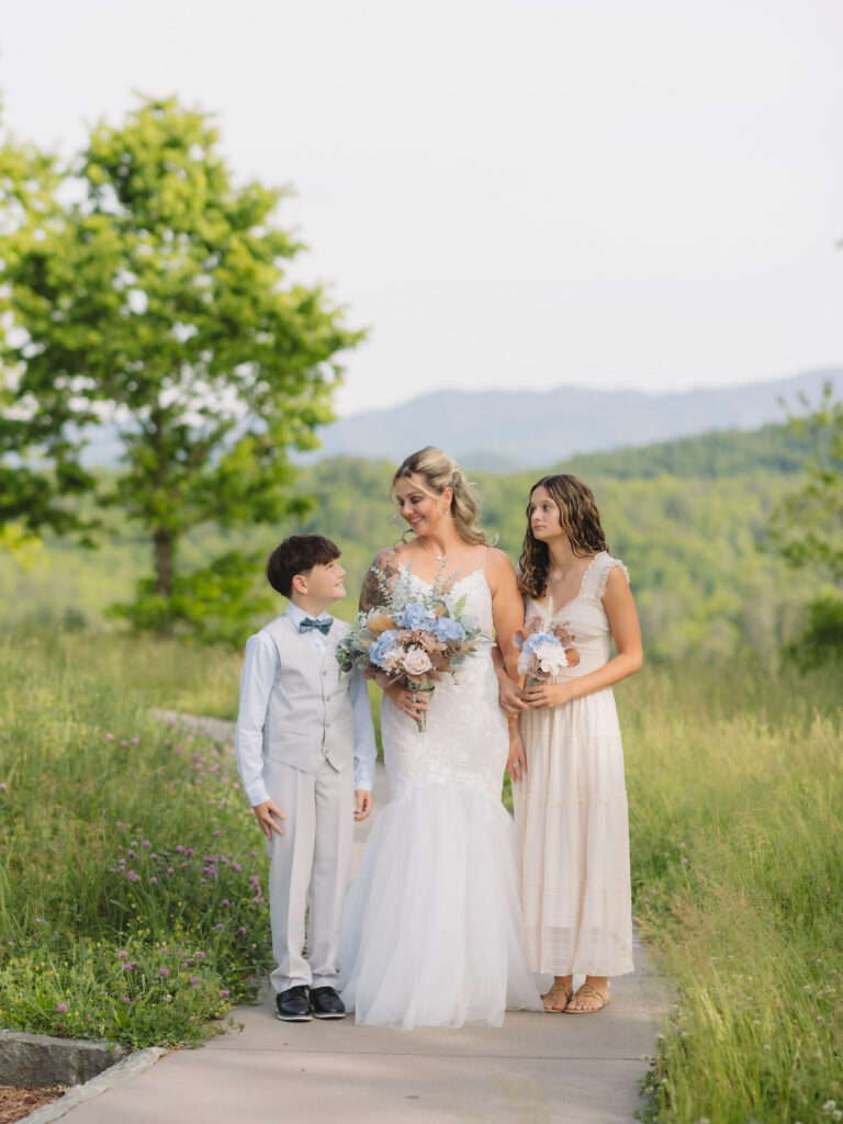 Bride poses with two young attendants holding pastel bouquets before a Smoky Mountain Ceremony surrounded by mountain views and summer greenery.