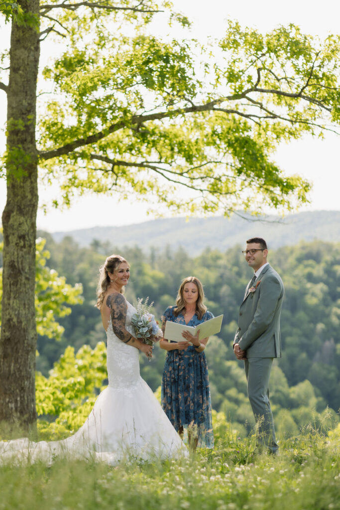 Bride and groom stand beneath a large tree with their officiant during a Smoky Mountain Ceremony overlooking layers of green mountain scenery.