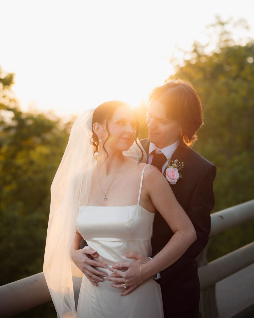 Bride and groom embrace in warm sunset light during their Foothills Parkway elopement, with her veil glowing against the wooded mountain backdrop.
