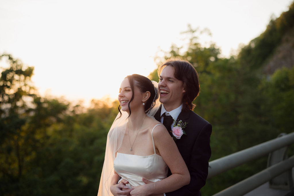 Newlyweds laugh together in warm sunset light during their Foothills Parkway elopement, with the bride in a satin gown and the groom wearing a pink rose boutonniere.