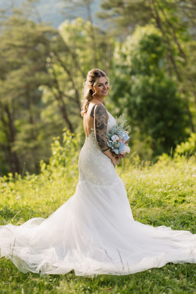 Bride poses in a lace wedding gown with a flowing train and pastel bouquet after her Smoky Mountain Ceremony surrounded by sunlit greenery.