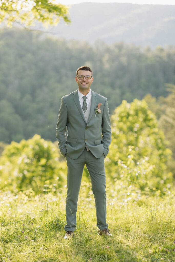 Groom in a sage green suit poses outdoors with a forested mountain backdrop during a Smoky Mountain wedding.