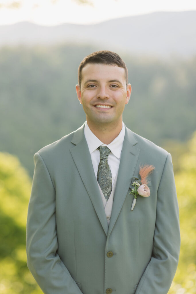 Groom in a sage green suit and floral boutonniere poses against soft mountain views for his Gatlinburg wedding photographer portrait.