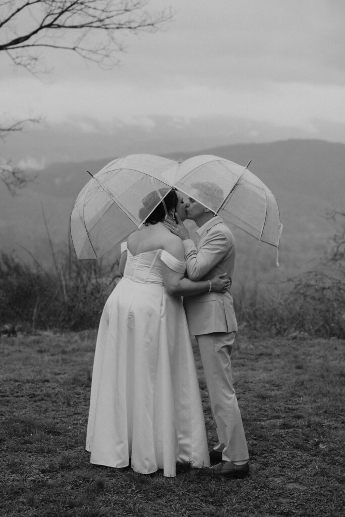 Newlyweds share a kiss beneath clear umbrellas in the rain while eloping to Gatlinburg at foothills parkway.