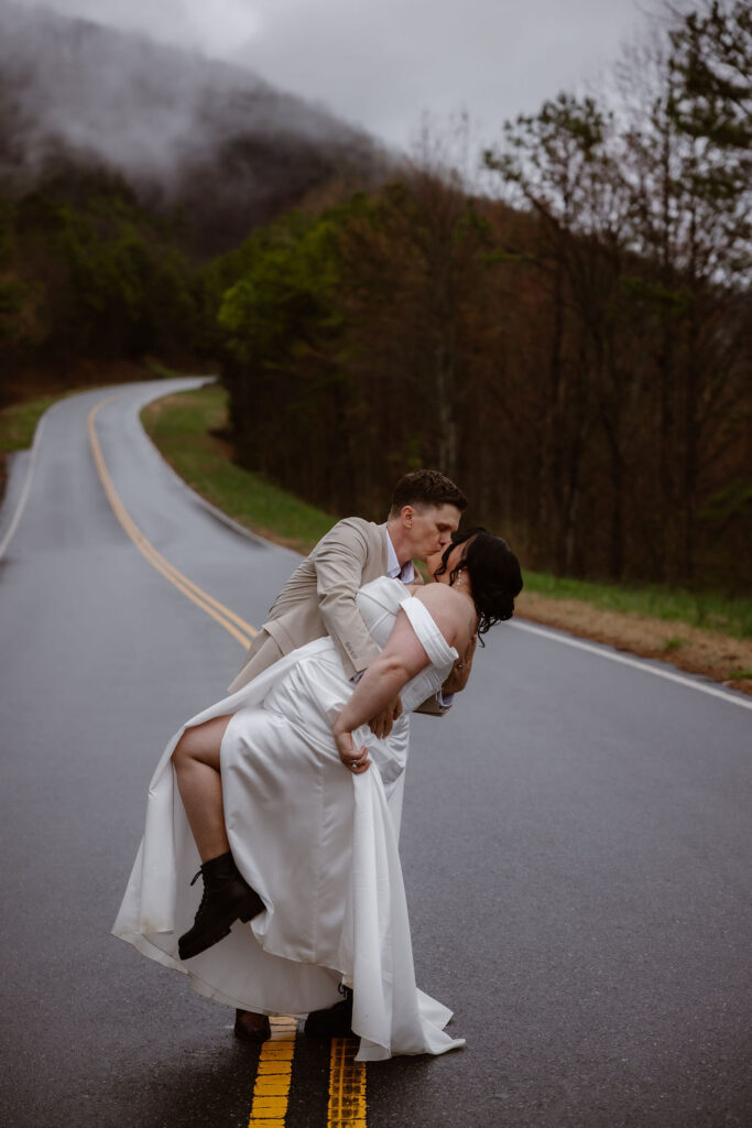 Newlyweds share a dramatic dip kiss on a mountain road while eloping to Gatlinburg at foothills parkway with misty Smoky Mountain views.