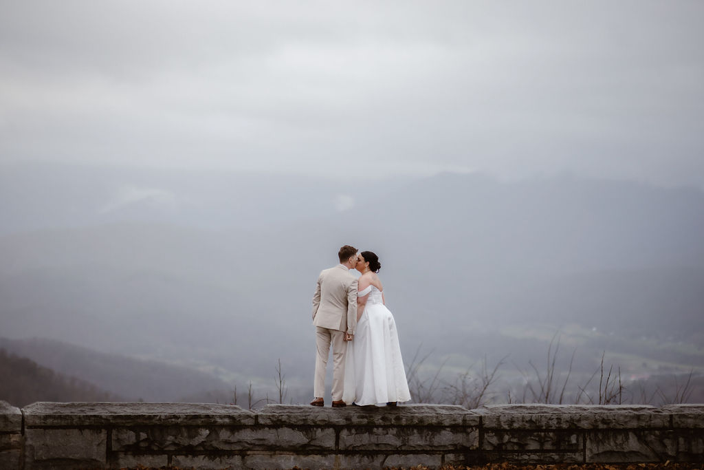 Couple sharing a quiet kiss on a scenic overlook while eloping to Gatlinburg, with misty mountain views along foothills parkway in the background.
