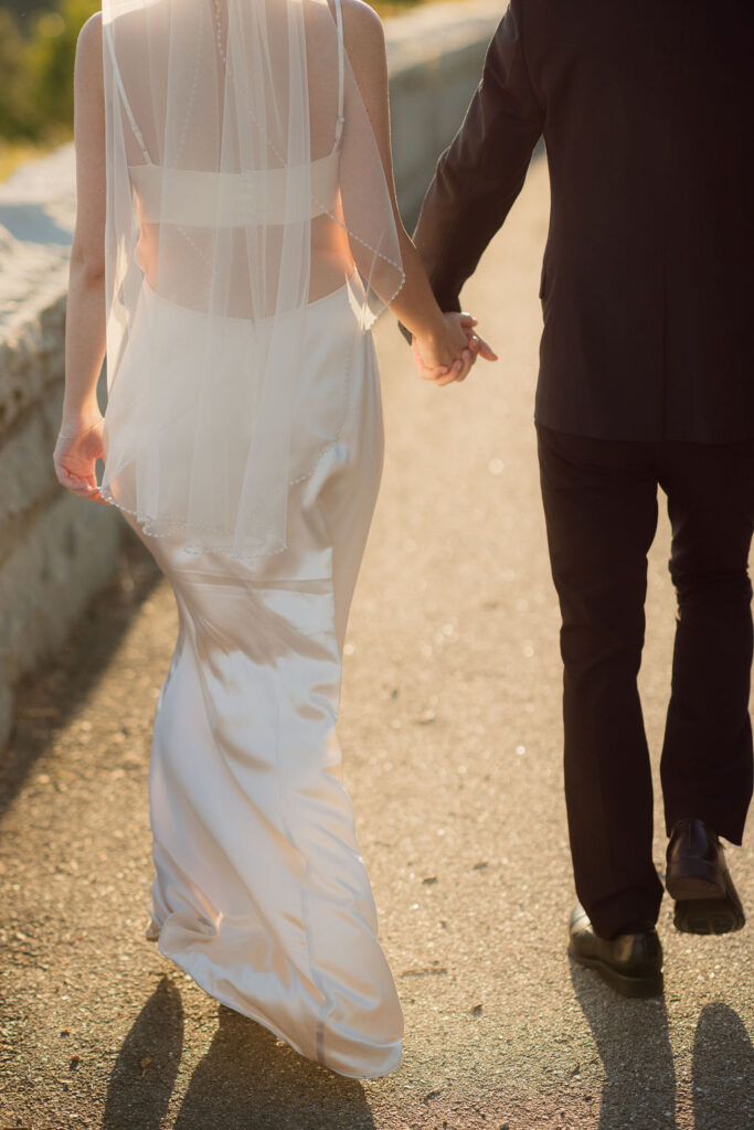 Bride and groom hold hands while walking along the sunlit Foothills Parkway, highlighting the bride’s satin gown and beaded veil.