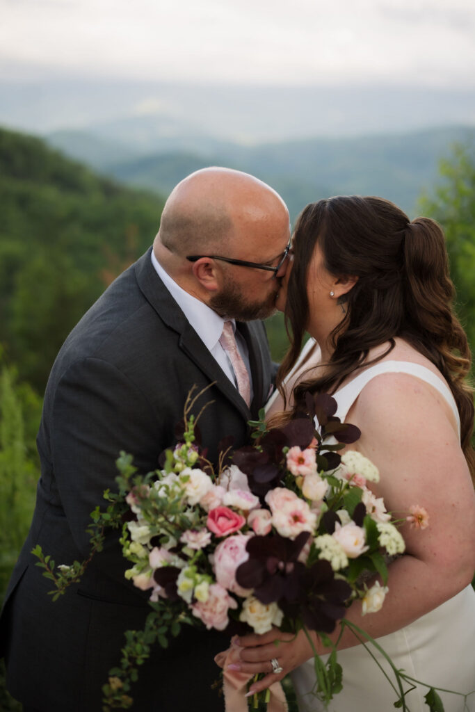 Newlyweds share a kiss with a vibrant bouquet and scenic mountain backdrop at Foothills Parkway while eloping to Gatlinburg.