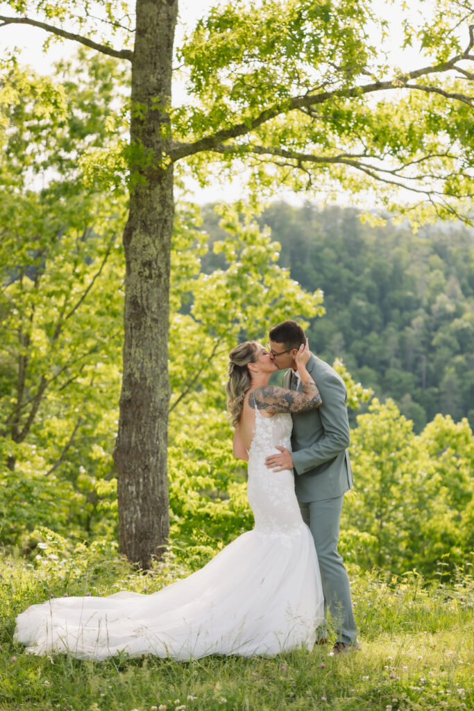 Bride and groom kiss beneath a large tree after their Smoky Mountain Ceremony, surrounded by sunlit forest and mountain views.