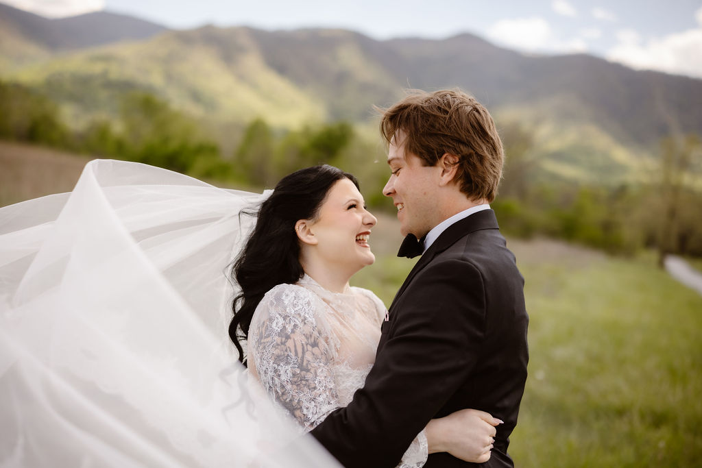 Bride and groom smile at each other beneath a flowing veil in Cades Cove, with the Great Smoky Mountains creating a scenic backdrop for eloping to Gatlinburg.