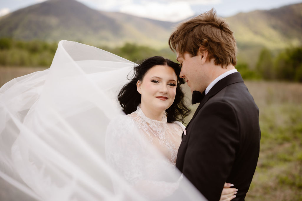 Bride and groom embrace in Cades Cove with the bride's veil flowing in the breeze and the Smoky Mountains in the background, capturing the beauty of eloping to Gatlinburg.