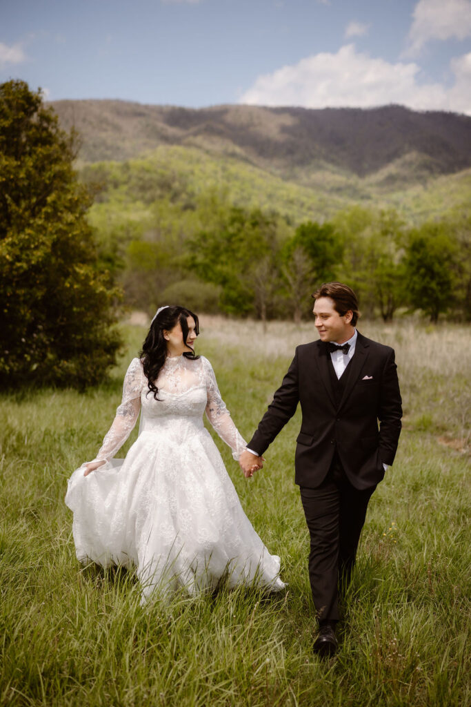 Bride and groom hold hands while walking through the Cades Cove meadow with the Smoky Mountains behind them, highlighting the beauty of eloping to Gatlinburg.