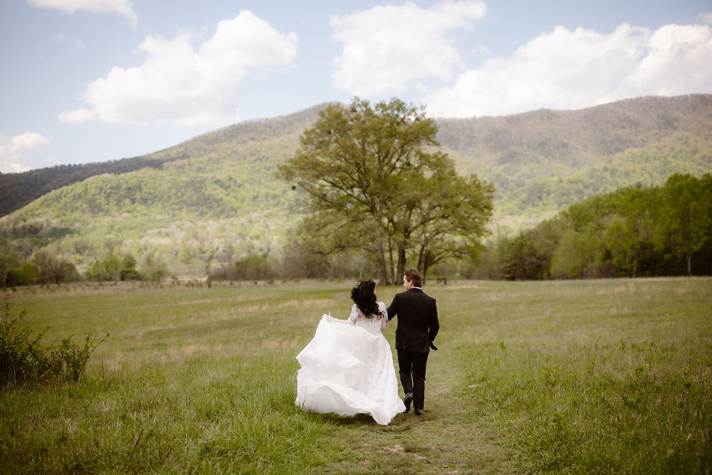Bride and groom walk hand in hand through a scenic meadow in Cades Cove, surrounded by mountain views, capturing the beauty of eloping to Gatlinburg.