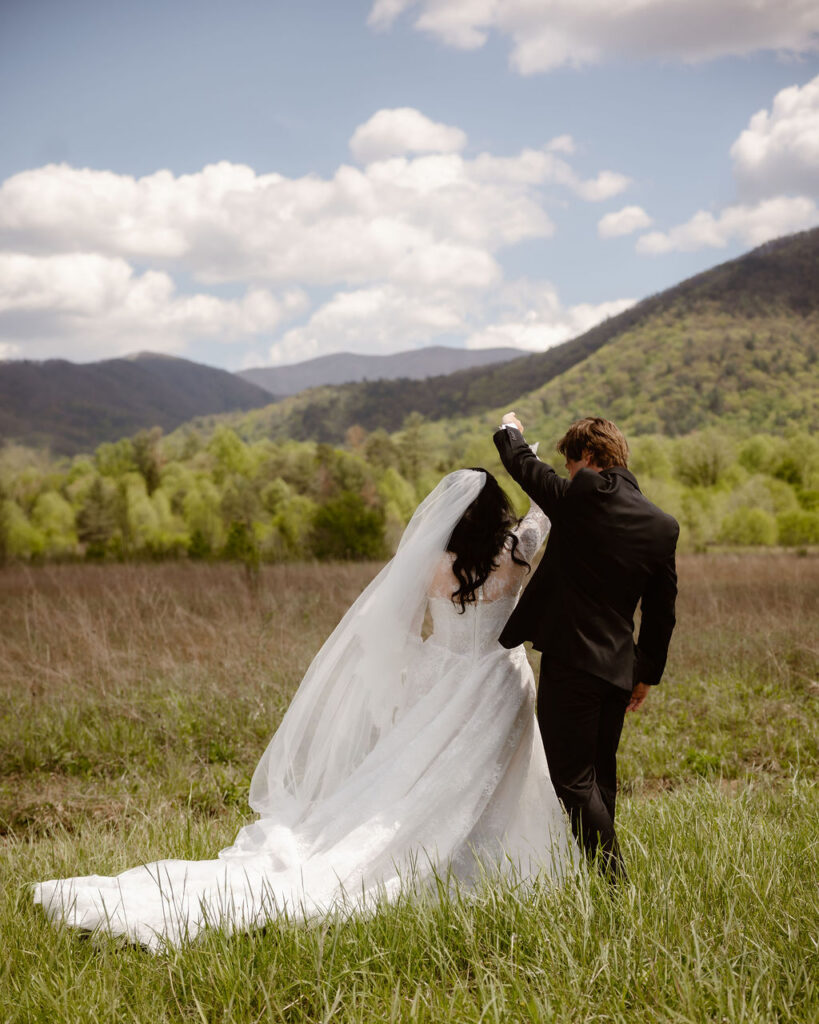 Bride and groom walk hand in hand through the meadow in Cades Cove with mountain views, celebrating the adventure of eloping to Gatlinburg.