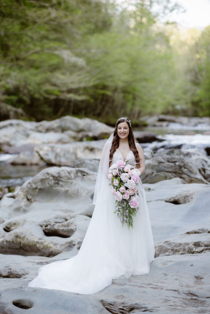Bride poses beside a rocky creek holding a cascading pink-and-white bouquet during her gatlinburg elopement portraits.