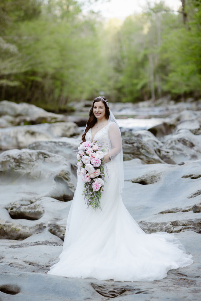 Bride poses with a cascading pink and white bouquet beside a mountain stream during a waterfall elopement.