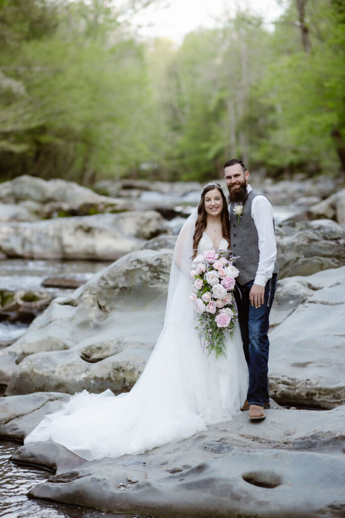 Bride and groom pose on smooth river rocks with a cascading pink-and-white bouquet during their greenbrier wedding portraits.