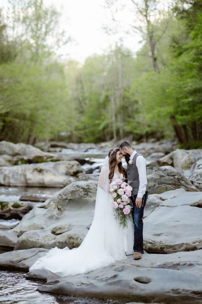 Bride and groom stand close together on river rocks surrounded by forest during their greenbrier wedding portraits.