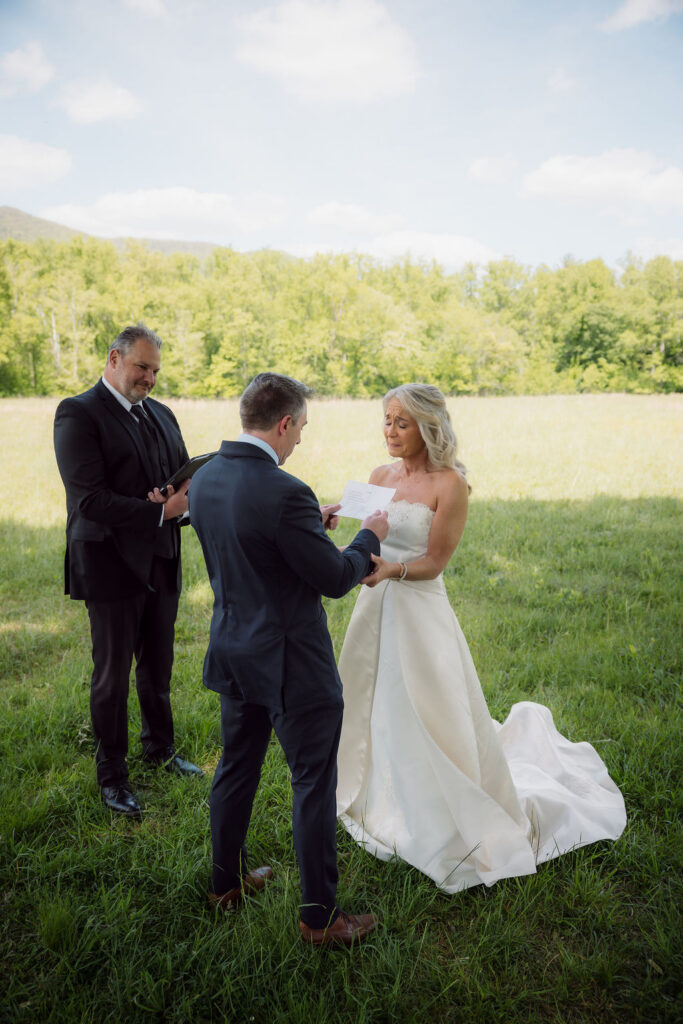 Groom reads vows to his bride as their officiant looks on during a Gatlinburg Elopement Ceremony in a sunlit mountain meadow.