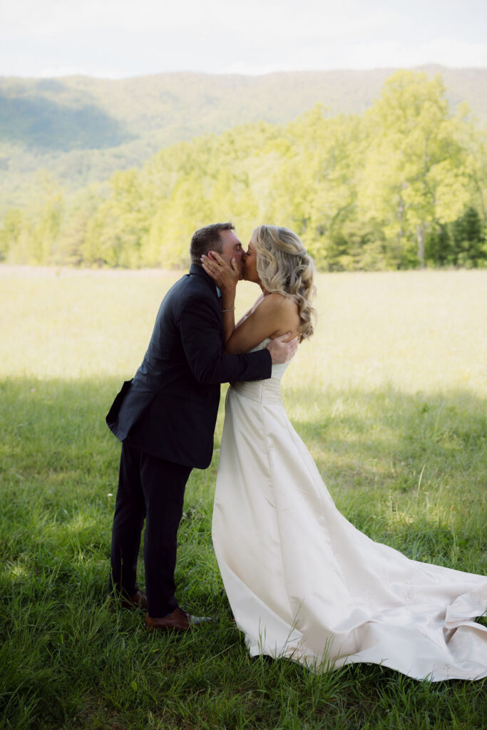 Newlyweds share a kiss in a sunlit meadow after their Gatlinburg Elopement Ceremony, with green Smoky Mountain ridges stretching across the background.