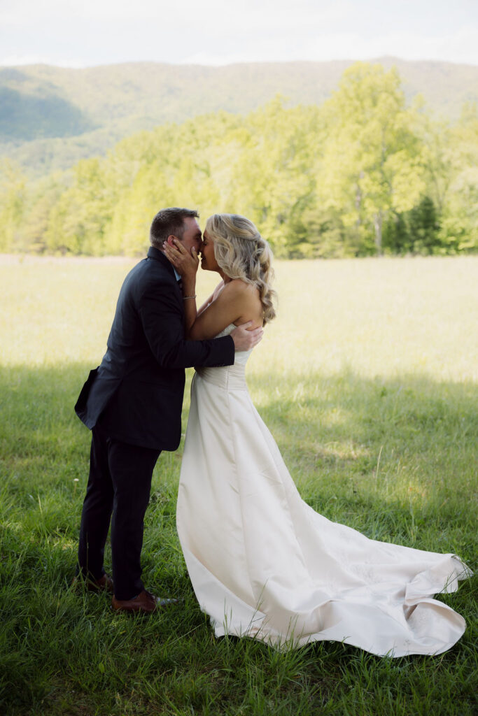 Newlyweds share their first kiss in a grassy mountain meadow during a cades cove elopement, with forested Smoky Mountain ridges behind them.