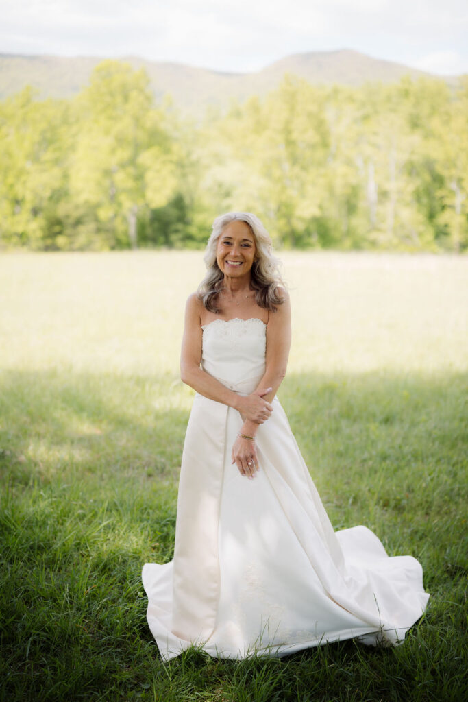 Bride poses in her wedding gown with sweeping mountain views behind her during a cades cove elopement, photographed in a quiet grassy meadow.