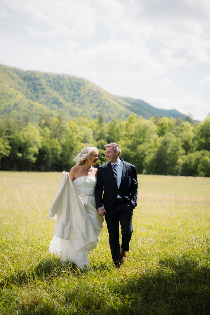 Bride and groom walk hand in hand through a sunlit meadow after eloping to gatlinburg, surrounded by sweeping mountain views and spring greenery.