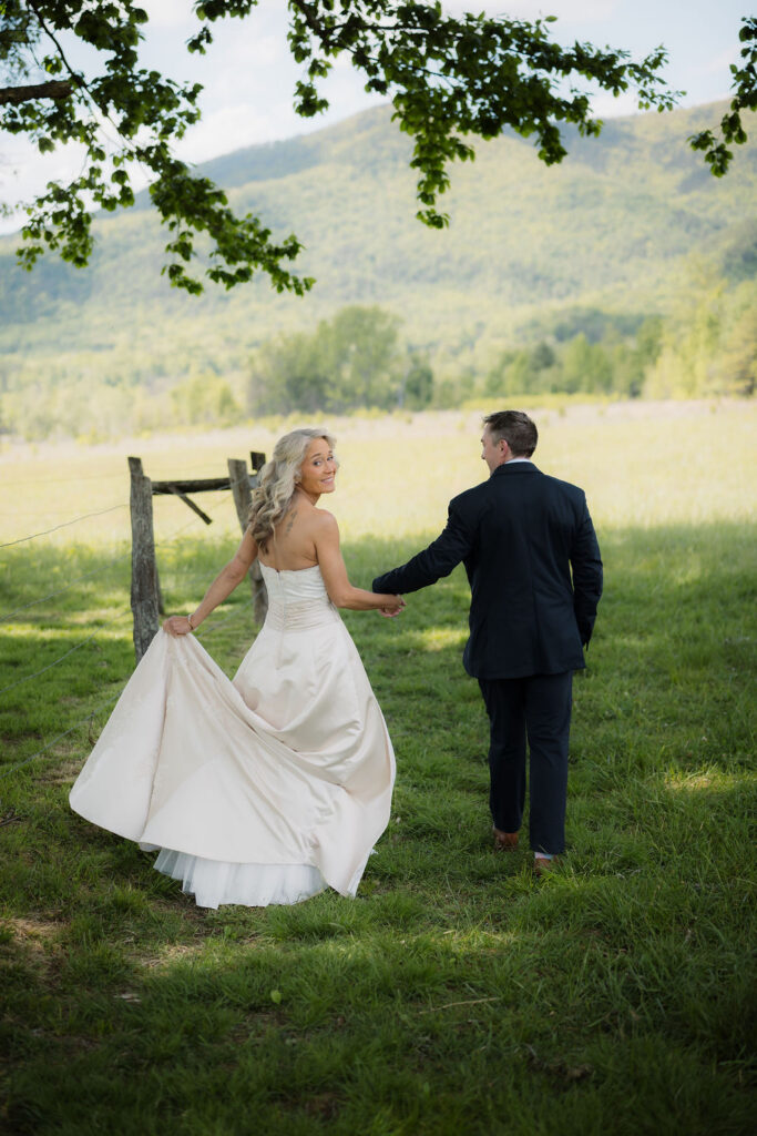 Bride looks back while walking hand in hand with her groom beneath leafy trees during their cades cove elopement, surrounded by open meadow and mountain views.