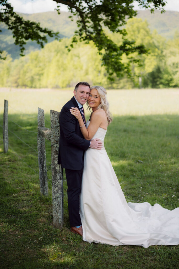 Newlyweds embrace beside a rustic fence during their cades cove elopement, framed by leafy trees, an open meadow, and Smoky Mountain views.