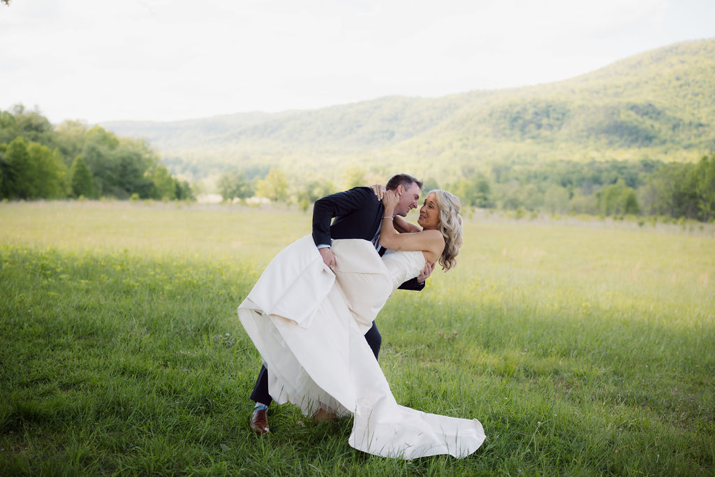 Groom playfully dips his bride in a grassy field while eloping to gatlinburg, captured by an elopement photographer with the Smoky Mountains behind them.