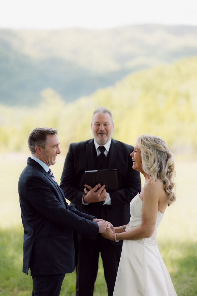 Bride and groom laugh while holding hands with their officiant behind them during an outdoor Gatlinburg Elopement Ceremony in the mountains.