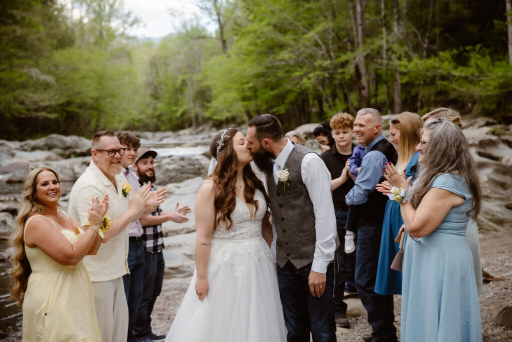 Newlyweds kiss as family and friends applaud beside the river during an intimate wedding in great smoky mountain national park.