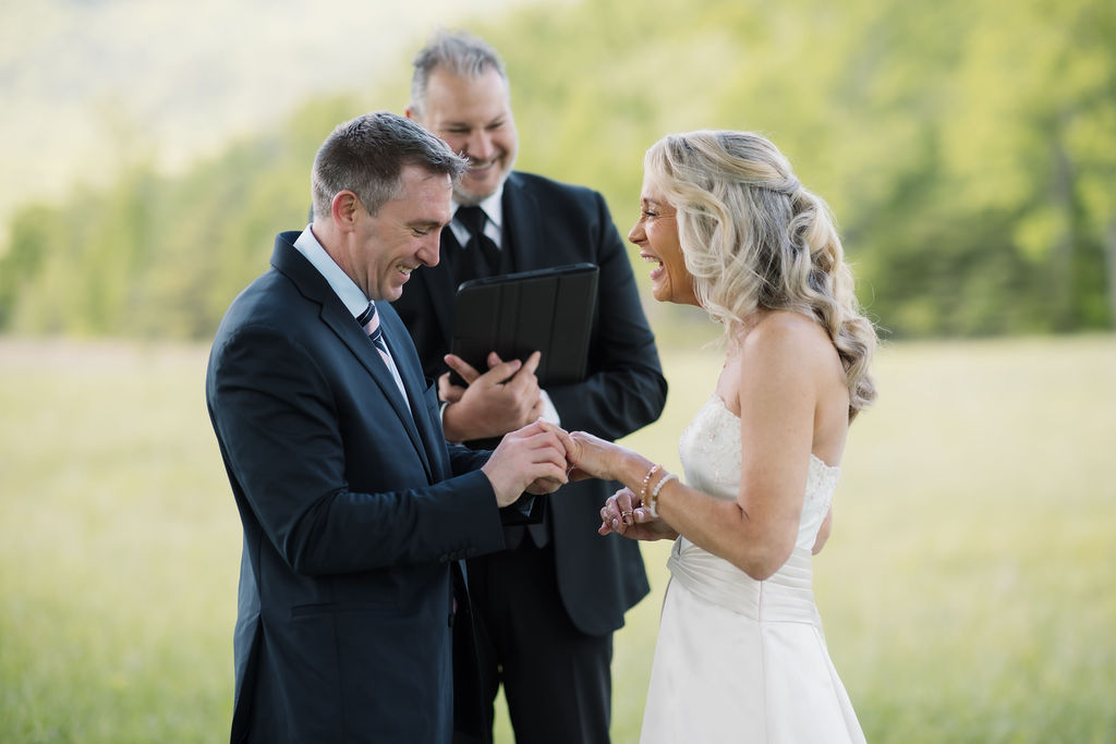 Groom places a wedding ring on his bride’s hand as they laugh with their officiant during an outdoor Gatlinburg Elopement Ceremony.