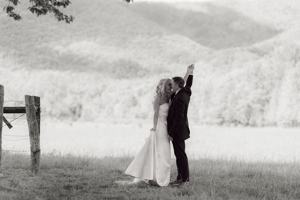 Black-and-white portrait of newlyweds kissing and raising their joined hands after eloping to gatlinburg, with a rustic fence and Smoky Mountains in the background.