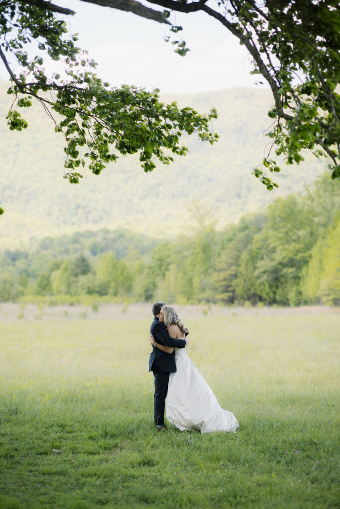 Newlyweds embrace in a sunlit mountain meadow during their cades cove elopement, surrounded by lush trees and sweeping Smoky Mountain views.