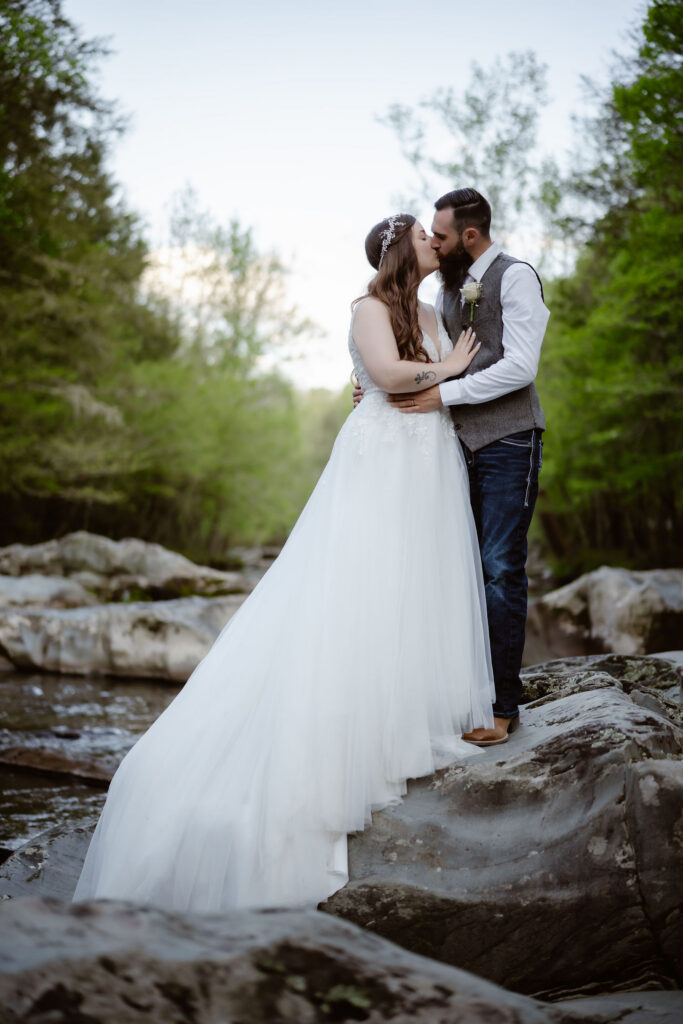 Bride and groom kiss on large river rocks surrounded by spring greenery while eloping to gatlinburg.