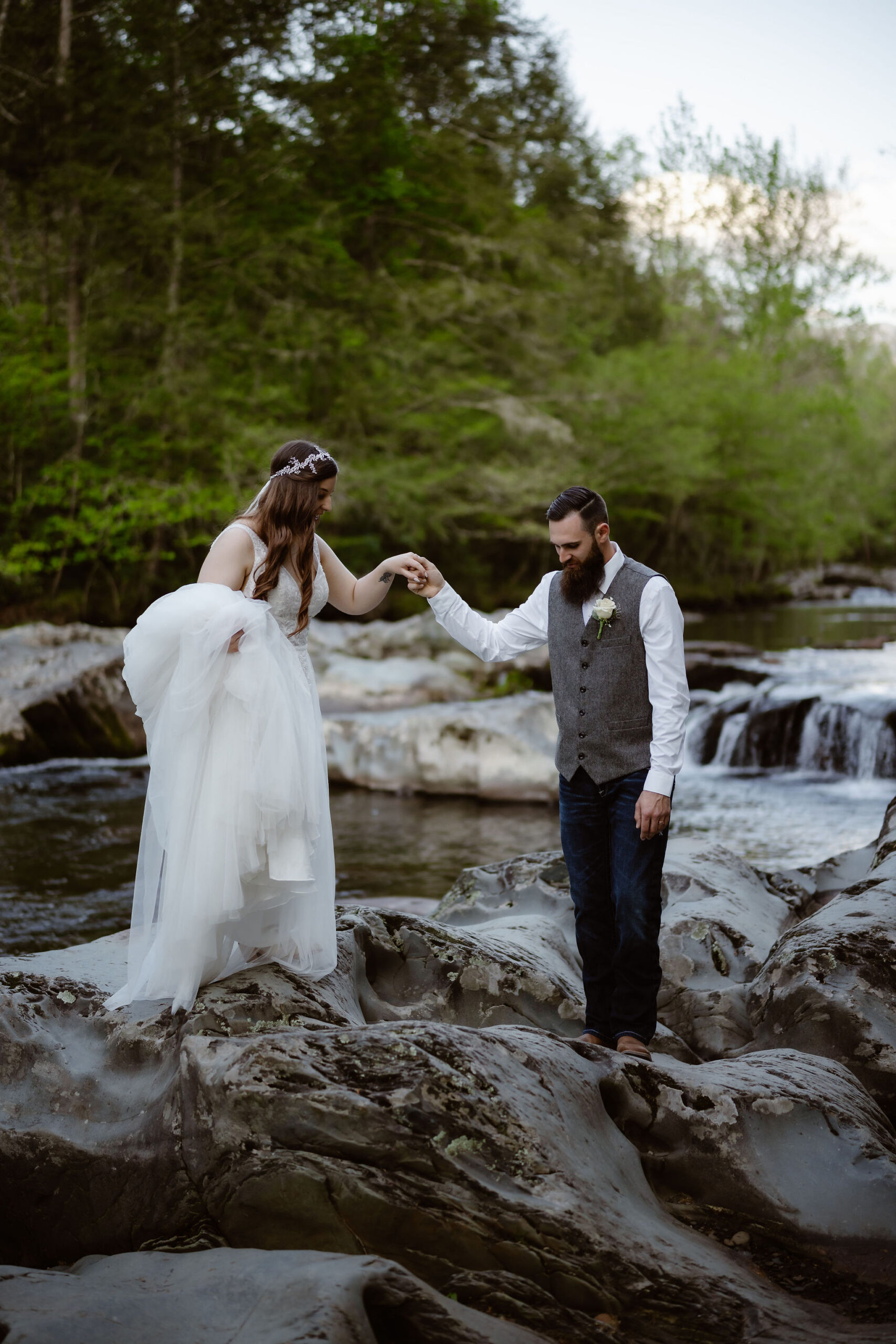 Groom holds the bride’s hand as she steps carefully across rocks beside a small cascade during their greenbrier elopement.