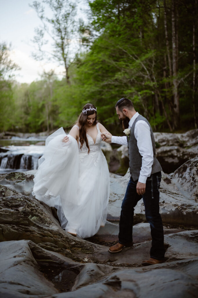 Groom holds the bride’s hand as she carefully crosses rocky terrain beside a forest stream during their waterfall elopement.