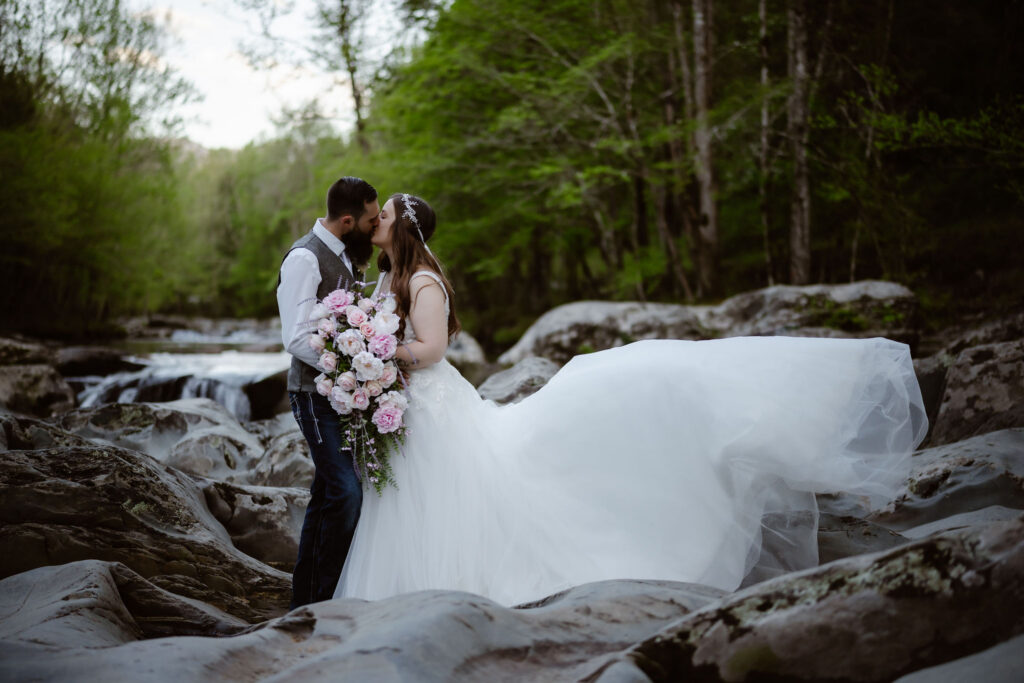 Bride and groom kiss beside a rocky forest stream, her flowing gown extended across the rocks during a gatlinburg elopement.