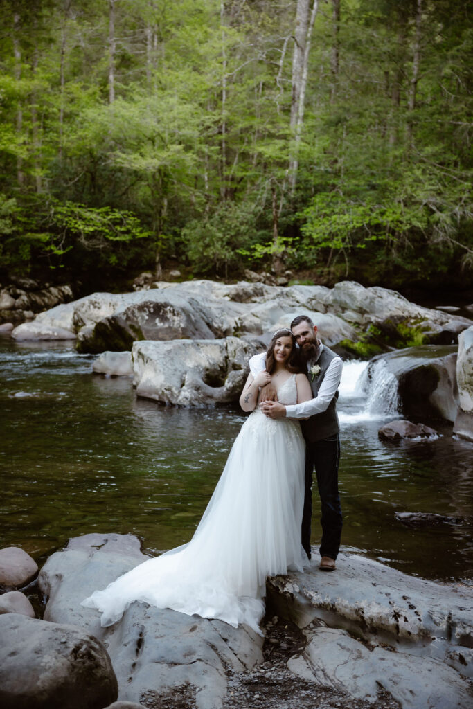 Bride and groom embrace on river rocks beside a small cascade in great smoky mountain national park, surrounded by dense spring forest.