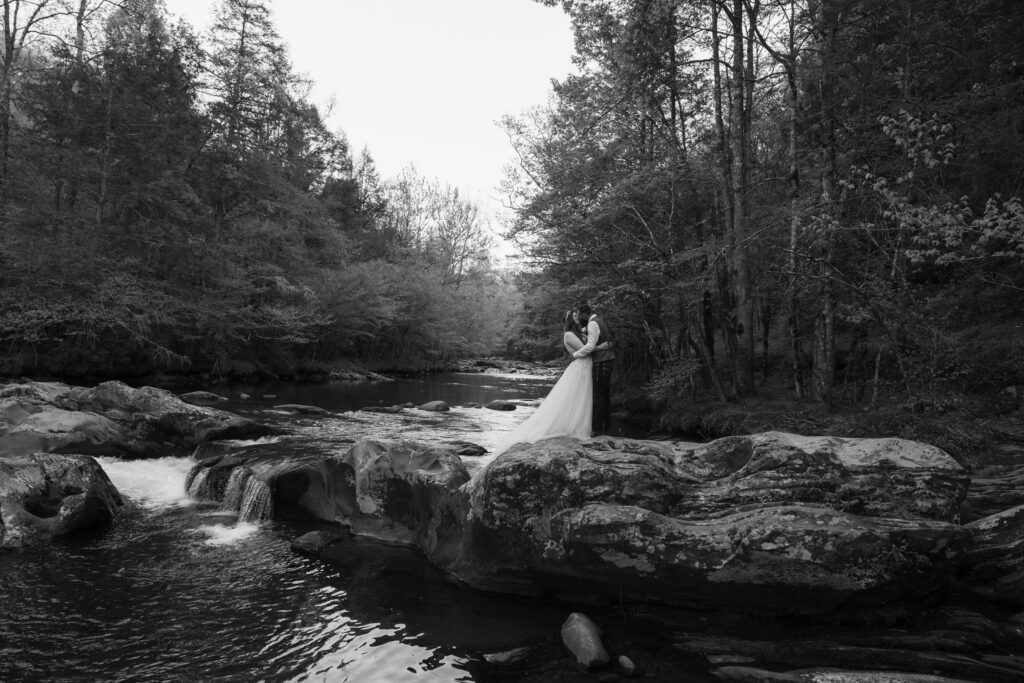 Black-and-white portrait of newlyweds kissing on river rocks beside rushing water and dense forest in great smoky mountain national park.