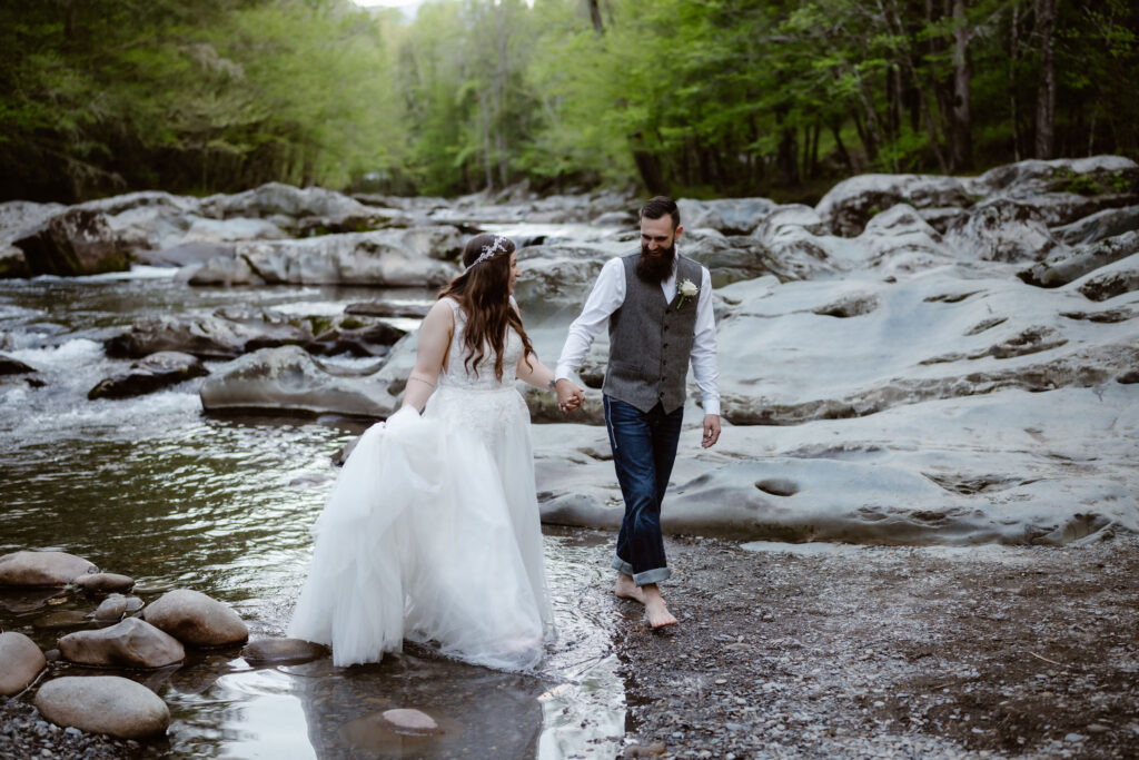 Newlyweds walk barefoot through a shallow mountain stream after their waterfall elopement, surrounded by rocks and fresh green forest.
