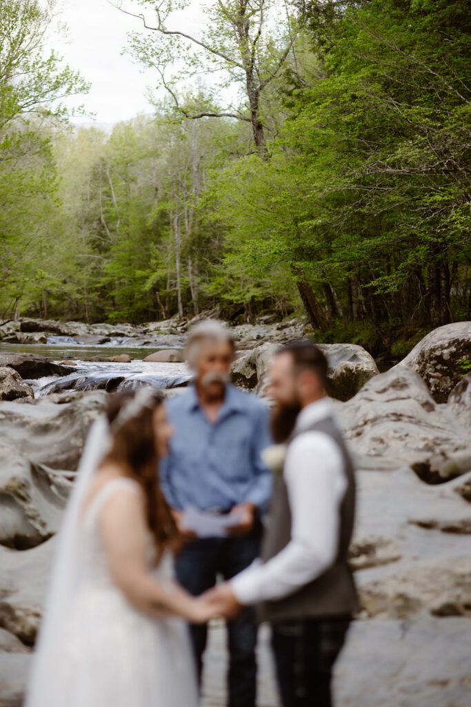 Bride and groom hold hands with their officiant beside a rocky mountain stream during an intimate greenbrier wedding ceremony.