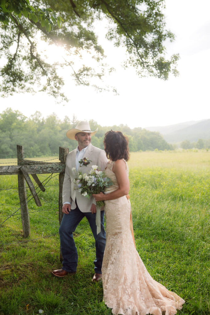 Bride and groom in a cowboy hat and champagne lace gown pose beside a rustic wooden fence in a sunlit meadow during their cades cove elopement.